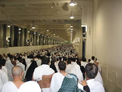 Large crowd of pilgrims in white Ihram walking through a long, brightly lit indoor walkway.