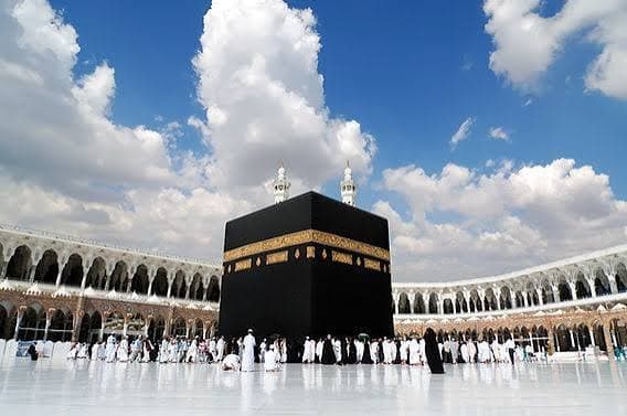 The Kaaba in Mecca surrounded by pilgrims in white ihram under a bright blue sky.
